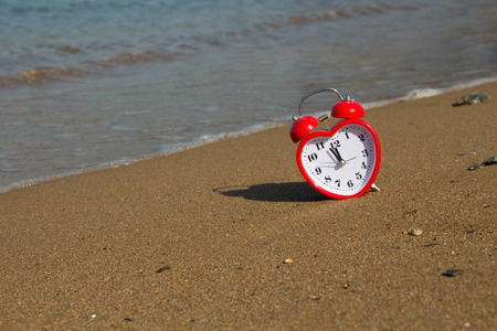Red clock in shape of heart on a sandy beach background.