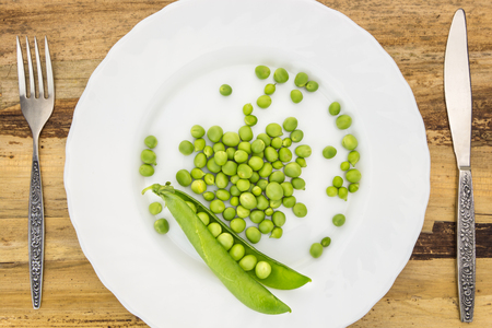 Fresh green peas on plate with fork and knife closeup, on wooden surfaceの写真素材