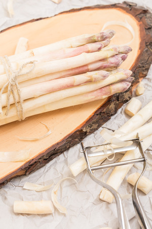 Bunch of white asparagus on  wood slice cutting board with metallic peeler,  on crumpled paperの写真素材