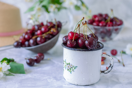 Close up Fresh cherry fruit in white mug, other dishes with berries and vase with jasmine flowers on the light marble table. Soft selective focus. Summer countryside concept.の写真素材