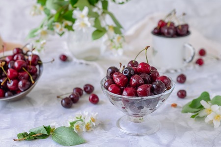 Fresh cherry fruit in glass vase, other dishes with berries and jar with jasmine and wildflowers on the light marble table. Soft selective focus. Summer countryside concept.の写真素材
