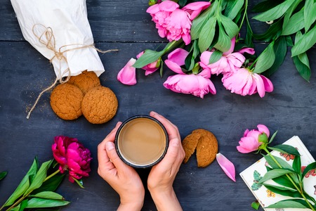 Top view close up female hands holding mug of coffee surrounded with cookies and peonies flowers. Coffee break concept. Selective focus, flatlay.の写真素材
