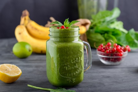 Healthy green spinach smoothie in a jar mug decorated with mint and red currant berries with ingredients on the black wooden table. Selective focus. Detox conceptの写真素材