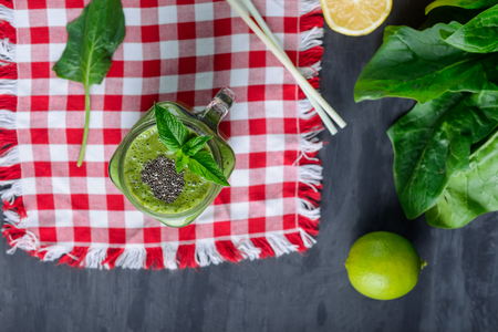 Top view Healthy green spinach smoothie in a jar mug decorated with mint and chia seeds with ingredients on the checkered red napkin on the black wooden table. Selective focus.の写真素材