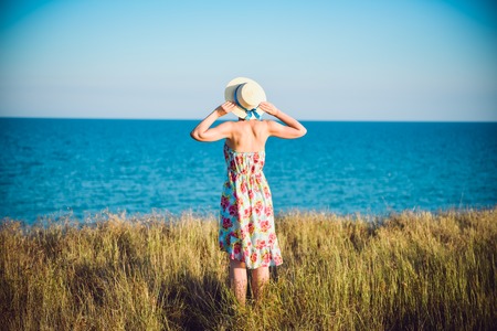 Summer portrait of young pretty woman in a straw hat and dress standing backwards in the grass and looking to the sea. Girl enjoy nature sea paradise and fresh air at wonderful summer vacation.の写真素材