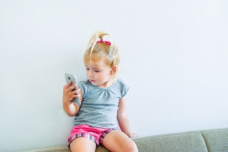 Close up Beautiful little baby girl holding and playing with smart phone sitting on the sofa on the white wall background. Children and technology concept. Selective focus. Space for text.の写真素材