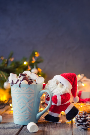 Decorative Santa Claus holding a cup of cocoa with marshmallow on the festive dark background with a Christmas tree, lights and decor on the old wooden boards. Selective focus. Text spaceの写真素材