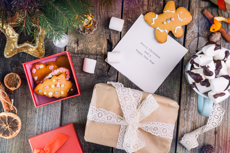 Christmas composition. Gifts, box with gingerbred cookies , cones, fir branches with decor, cup of cocoa with murshmellows and greeting card with wishes on wooden vintage background. Selective focusの写真素材