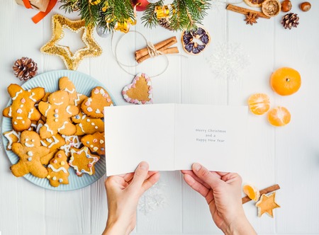 Top view female hands holding greeting card with wishes under white wooden table with gingerbred cookies, christmas tree branches with decor, spices. Selective focus. Text space.の写真素材