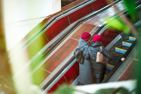 Back view two girls moving up on escalator in subway. Escalator in metro. Moving up staircase. electric escalator. View through the greenery. Selective soft focus.の写真素材