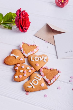 Homemade cookies in shape of heart with I Love you words as gift for lover on Valentine's day. White wooden table with greeting card, rose and decor. Festive gift concept. Selective focus. Copy space.の写真素材