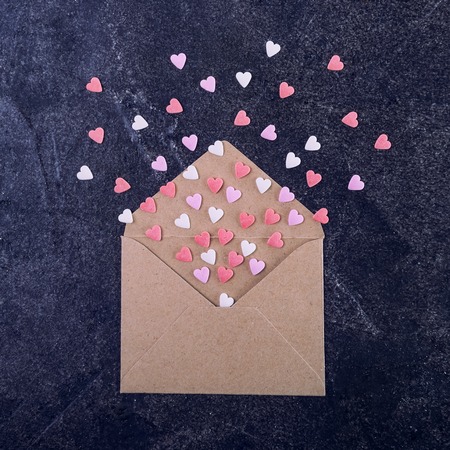 Pink, red and white sweets sugar candy hearts fly out of craft paper envelope on the dark stone background . Valentine day concept. Gift for lover. Selective focus. Space for text. Square card.の写真素材