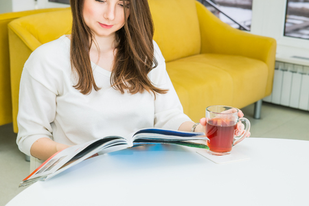 Close up oung girl sitting at the table in modern waiting hall with bright yellow sofas, reading a magazine and drinking tea. Hospitality, medicine, business concept. Selective focus. Space for textの写真素材