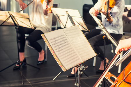 Selective Focus on Music note sheets on stand with background of playing cellists and violinists band on event in bright day sunlight. Commercial music. Service at business meeting, party, weddingsの写真素材