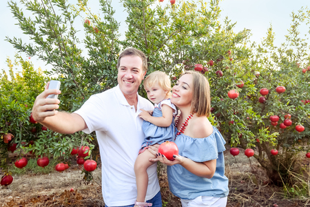 Portrait of smiling parents with baby girl daughter doing selfie photo with smart phone in the pomegrate fruit garden on the sunset. Happy family vacation. Sunset light. Selective focus. Copy spaceの写真素材