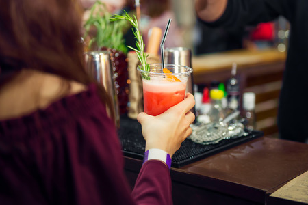 Backview girl hands holding plastic glass with red coctail with bar counter on the background. Leisure and lifestyle, drinking cocktails, nightlife and party event concept. Sective focus. Copy spaceの写真素材