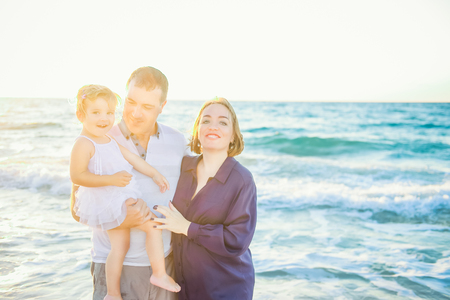 Happy family of three - pregnant mother, father and daughter embracing, laughhing and having fun walking on the beach. Family vacation, travel concept. Backlight, soft selective focus. Copy spaceの写真素材