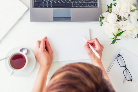 Workspace with girls hands writing in notebook with empty blank, laptop keyboard, cup of tea, glasses, white peony on woodden table. Top view feminine office desk. Freelancer working place.の写真素材