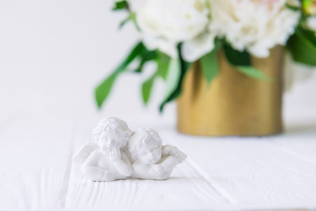 Close up statuette of two antique little lovely angels of the gypsum on the white wooden table with bouquet of white peonies in vintage bowl. Love background. Copy space, selective focusの写真素材