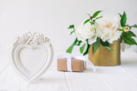 Heart shaped photoframe with plaster flowers,gift box with ribbon and vintage bowl with white peonies bouquet on the wooden table. Love background. Greeting card. Copy space, selective focusの写真素材