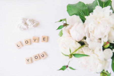 Flat lay Love you lettering spelled in wooden blocks with statuette of two antique lovely angels with blurred white peonies on light background. Love words. Valentine's day. Top view. Copy spaceの写真素材