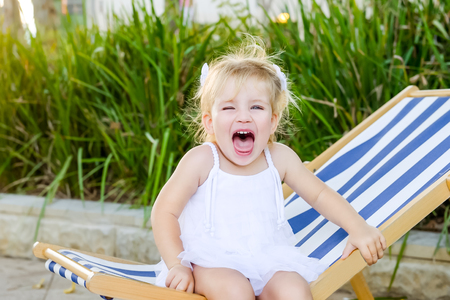 Close up portrait of cute emotional blondy toddler girl in white dress sitting on the deckchair and yelling. City park recreation area. Selective focus, copy spaceの写真素材