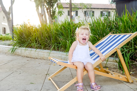 Portrait of cute emotional blondy toddler girl in white dress sitting on the deckchair and yelling. City park recreation area. Selective focus, copy spaceの写真素材