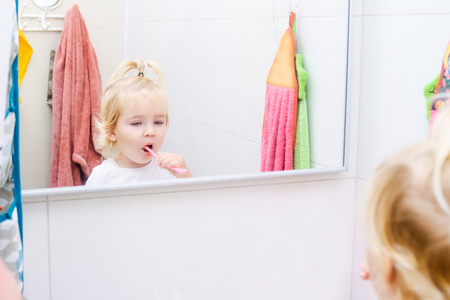 Close up little toddler daughter brushing her teeth and looking in the mirror in the bathroom. Reflection in the mirror. Family health care, everyday habits. Selective focus, copy spaceの写真素材