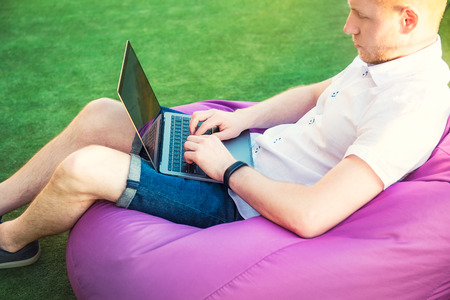 Young man in casual clothes sitting on pouf, working on laptop pc computer in park on green lawn outdoors. Mobile Office, Comfortable work place concept. Freelancer. Soft selective focus. Copy spaceの写真素材