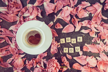Flatlay cup of coffee with heart shape pattern and message I love you spelled in wooden blocks with pink peony flower petals on dark background.の写真素材