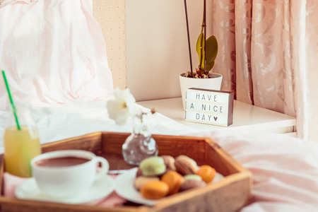 Focused Have a nice day text on lighted box on the bedside table and blurred wooden tray with coffee and macaroons breakfast on bed. Good morning mood. Hospitality, care, service concept. Copy spaceの写真素材