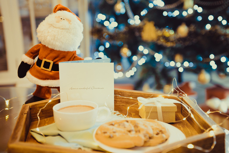 Decorative Santa standing near tray with cocoa, cookies, giftbox and postcard with xmas tree background. Christmas and New Year composition. Festive breakfast. Soft selective focus. Vintage toningの写真素材