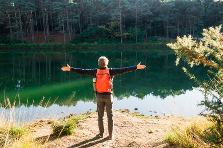 Back view young tourist man with backpack arms raised up to sky, celebrating freedom standing near forest lake. Exploring the world. Walking adventure. Selective focus, copy space. Blue poole, Ukの写真素材