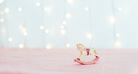 Vintage Christmas tree toy porcelain figurine of a rocking horse standing on the pink tablecloth against the background of blurred lights. Festive layout. Wide banner. Soft selective focus. Copy space.の写真素材