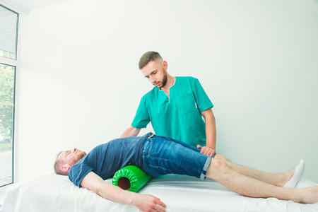 Man at the physiotherapy doing physical exercises with his therapist, they using a massage roll. A chiropractor treats patient's loins spine in medical office. Neurology, Osteopathy, chiropracticの写真素材