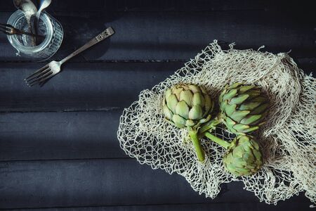 Top view Fresh green artichokes with braided rope and kitchen stuff on the black wooden table lightened by morning window light. Farmer harvest, local market. Selective focus, Copy space. Flat layの写真素材