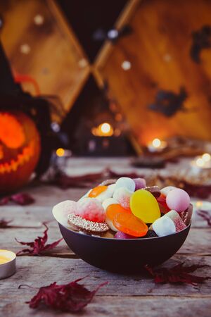 Close up bowl with candies and carved pumpkin in witch hat with paper silhouettes of bats, castle, ghosts on wooden background. Spooky holiday symbols. Trick or treat concept. Copy spaceの写真素材