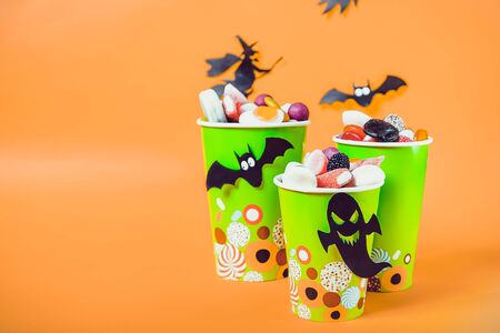 Halloween trick or treat sweets. Paper cups with colourful candies inside and paper silhouettes of bats, ghosts and witch on bright orange background.の写真素材