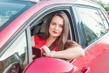 Young business woman driver in red suit holding and looking on her cell smart phone while sitting behind the wheel red car. Selective focus, copy spaceの写真素材