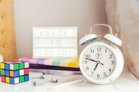 Close up focused white alarm clock with blurred Back to school message hashtag on lightened box, notebooks, rubik's cube and stationery on the bedside table. Conceptual Background. Copy spaceの写真素材