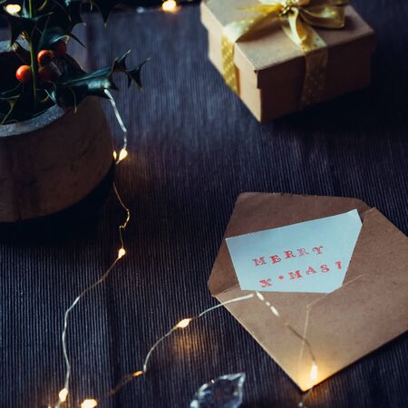 Festive composition of Merry Christmas card in craft envelope, gift box, berry holly ilex aquifolium...