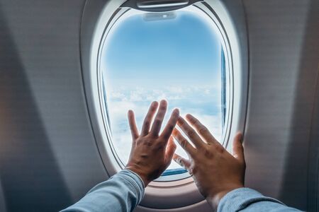 Man's and woman's hands inside touching window of airplane with clouds and blue sky background ....