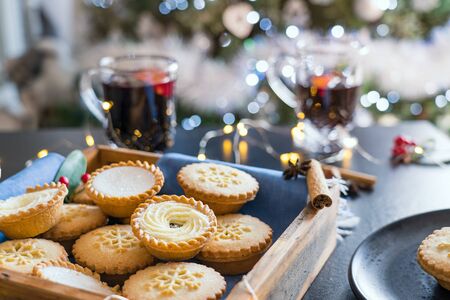 Warm cosy composition of traditional english festive pastry mince pies in wooden tray with blurred background of mulled wine drinks, lights garland and christmas tree on background. Close up.の写真素材