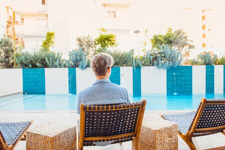 Back view young caucasian man in blue shirt relaxing on chair near swimming pool watching running water and plant flowebed. Summer, travel, vacation and holiday concept. Slow lifestyle. Copy spaceの写真素材