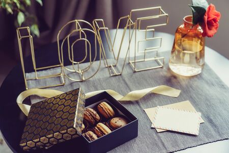 Festive box with chocolate macaroons cakes, rose flower in vase, postcard and wire word Love on gray...