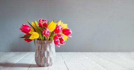 Fresh spring yellow and pink tulips bouquet in blue vase standing on black wooden table with gray background. Festive flowers for mother's or women's day. Mockup for greeting wide banner. Copy spaceの写真素材