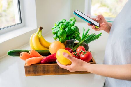 Online home delivery of fresh vegetables and fruits. Woman holding phone in one hand and lemon in other from wooden box with bio vegetables standing on white kitchen table. Local farmer healthy foodの写真素材