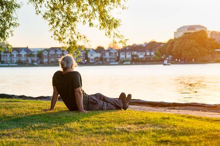 Back view gray hair mature man sitting on the green grass at the bank of park's lake and enjoying the sunset. A simple pleasure for mental health. Nature relaxation. Selective focus. Copy spaceの写真素材