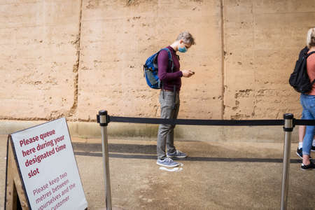 Young caucasian man wear face mask using his mobile phone while waiting social distance queue to...