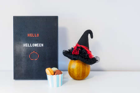 Minimalist Halloween decor - letter board with Hello Halloween message, witch hat on decorative pumpkin and cup with sweet macaroons on white background. Festive concept. Copy space.の写真素材